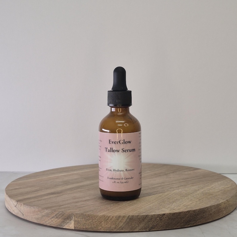 Op de afbeelding: Een bruine glazen fles EverGlow Tallow Serum met een zwarte druppelaar. Het etiket is roze en wit, met de woorden "Firm, Hydrate, Restore" en "Frankincense & Lavender". De fles staat op een houten, ronde schaal.