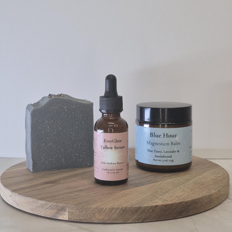 Op de afbeelding: Een houten dienblad met een grijze zeep, een bruine glazen fles EverGlow Tallow Serum en een pot Blue Hour Magnesium Balm. De serumfles heeft een druppelaar en de balsempot heeft een zwart deksel. De zeep heeft lavendelbloesem.
