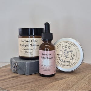 Op de afbeelding: Een verzameling huidverzorgingsproducten, waaronder een pot "Morning Glow Whipped Tallow", een fles "Ever-Glow Tallow Serum" en een blikje "Nourishing Salve". Gepresenteerd op een houten plank.