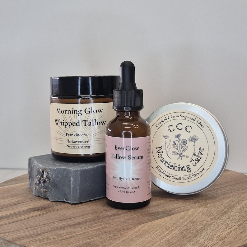 Op de afbeelding: Een verzameling huidverzorgingsproducten, waaronder een pot "Morning Glow Whipped Tallow", een fles "Ever-Glow Tallow Serum" en een blikje "Nourishing Salve". Gepresenteerd op een houten plank.