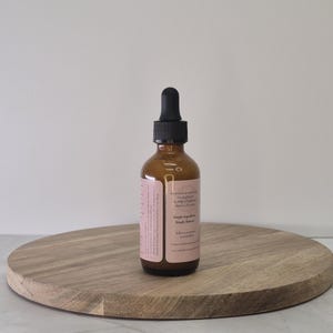 Op de afbeelding: Een bruine glazen fles met een zwarte druppelkap, rustend op een ronde houten plank. De fles heeft een roze etiket met tekst. De achtergrond is een effen witte muur.