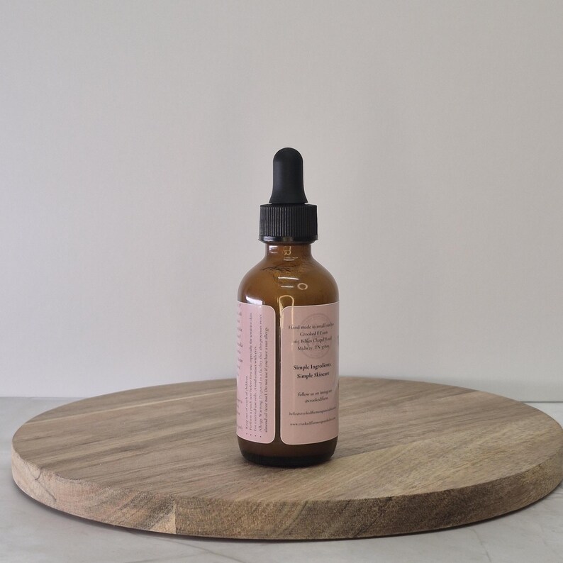 Op de afbeelding: Een bruine glazen fles met een zwarte druppelkap, rustend op een ronde houten plank. De fles heeft een roze etiket met tekst. De achtergrond is een effen witte muur.