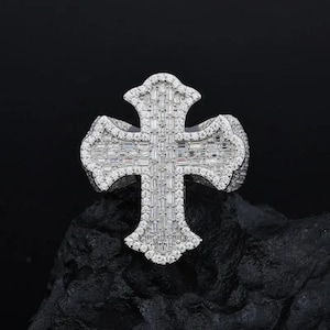 May include: A silver-toned cross ring with a detailed design. The cross is encrusted with numerous small, rectangular, and round-cut clear stones, creating a sparkling effect. The ring is set against a dark background.