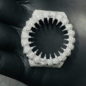 Könnte beinhalten: Eine diamantbesetzte Lünette für eine Uhr mit einem einzigartigen Design. Die Lünette ist sechseckig, mit einer strukturierten Oberfläche und einer Reihe spitzer, dreieckiger Elemente am Innenrand. Das Wort "icecraft" ist sichtbar.