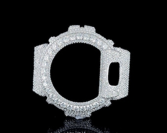 Könnte beinhalten: Eine silberfarbene Lünette für eine Uhr, besetzt mit zahlreichen kleinen, funkelnden Edelsteinen. Die Lünette hat ein kreisförmiges Design mit einer rechteckigen Öffnung an der Seite. Das Uhrenzubehör ist vor einem schwarzen Hintergrund platziert, mit einer Reflexion darunter.