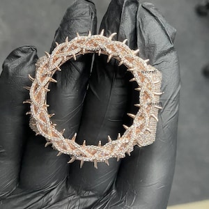 May include: A rose gold-toned bracelet in the shape of a crown of thorns, encrusted with small, sparkling white gemstones. The bracelet features sharp, pointed thorns evenly spaced around the band. The word "icecraft" is visible on the bracelet.