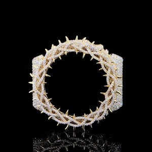 May include: A gold-toned bracelet with a thorn design, encrusted with sparkling crystals. The bracelet features a circular shape with sharp, pointed thorns evenly spaced around the outer edge. The jewelry is set against a black background.