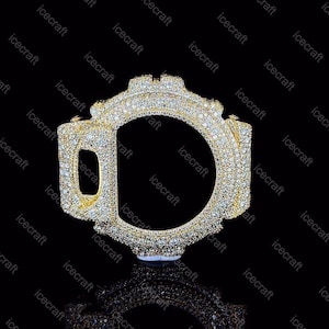 Puede incluir: Un bisel de reloj en tono dorado completamente incrustado con numerosas piedras pequeñas, claras y brillantes. El accesorio presenta un diseño robusto y multifacético con una abertura circular central y un recorte lateral rectangular.