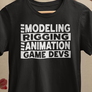 May include: Black t-shirt with white text stacked vertically: MODELING, RIGGING, ANIMATION, GAME DEVS. The shirt is made of a soft material and has a crew neck. The shirt is displayed on a wooden hanger.