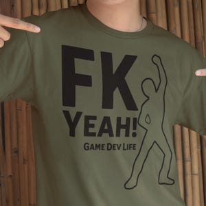 May include: Olive green t-shirt with the text "FK YEAH!" in large black letters, a black outline of a person celebrating, and the words "GAME DEV LIFE" below. The shirt is being pointed at by two hands.