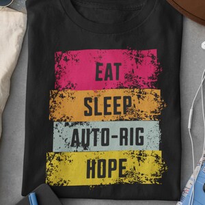 May include: Black t-shirt with the words "EAT SLEEP AUTO-RIG HOPE" in a distressed, colorful design. The text is stacked in horizontal bands of pink, orange, blue, and yellow. The shirt is made of a soft material.