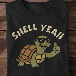 Peut inclure: T-shirt noir avec l'inscription "SHELL YEAH" au-dessus d'une tortue de dessin animé portant des lunettes de soleil et faisant un signe de pouce. La tortue a une carapace marron et verte.