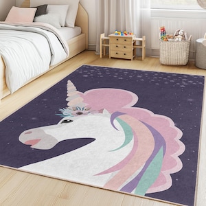 Può includere: Un tappeto rettangolare con un design a unicorno. Il tappeto ha uno sfondo blu scuro con una testa di unicorno bianca, una criniera rosa e una corona floreale. Il tappeto è posto nella camera da letto di un bambino.