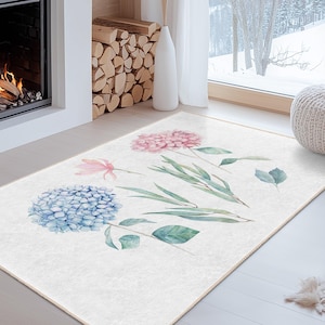 May include: A rectangular rug with a watercolour floral design. The rug has a light cream background with blue and pink hydrangea blooms, green leaves, and a pink tulip. The rug is placed on a light wood floor.