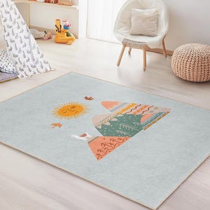 Könnte beinhalten: Rechteckiger Teppich mit hellblauem Hintergrund und einem farbenfrohen Berg- und Sonnendesign. Der Berg weist orange, grüne und rosa geometrische Muster auf. Eine gelbe Sonne und Vögel sind ebenfalls abgebildet.