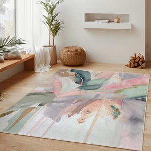 May include: A rectangular area rug with an abstract watercolor design in shades of pink, green, gray, and peach. The rug is placed on a hardwood floor in a living room setting, with a woven pendant light and plants.
