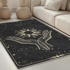 May include: A rectangular black area rug with a celestial design. The rug features a sun, hands, stars, and moon phases in a cream color. The rug is placed in a living room setting with a cream-colored sofa and decorative vases.