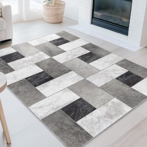 May include: A rectangular area rug with a geometric pattern in shades of gray, white, and black. The rug features a repeating design of rectangular shapes, creating a modern and stylish look. The rug is placed on a light-colored floor in a living room setting.