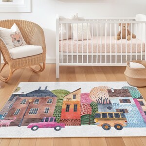 May include: A rectangular rug with a watercolour illustration of a town scene, including buildings, trees, a pink car, and a yellow school bus. The rug is mainly white with colourful details, suitable for a nursery or children's room.