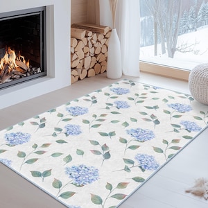 May include: A rectangular rug with a floral design. It displays light blue hydrangea flowers and green leaves against a cream backdrop. The rug is positioned in front of a fireplace and a large window.