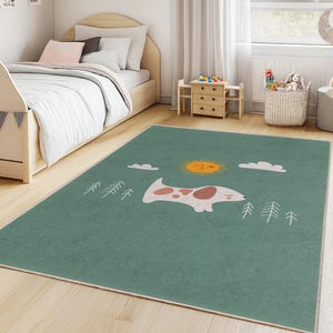 Könnte beinhalten: Ein grüner rechteckiger Teppich mit einem skurrilen Design in einem Kinderzimmer. Der Teppich zeigt eine Sonne, Wolken, Bäume und eine Illustration eines weißen und braunen Tieres. Der Teppich liegt auf einem hellen Holzboden.