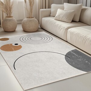 Può includere: Un tappeto rettangolare color crema con forme geometriche astratte in nero, marrone e grigio. Il tappeto presenta un grande semicerchio, cerchi e forme ovali. Il tappeto si trova in un soggiorno con un divano e vasi decorativi.