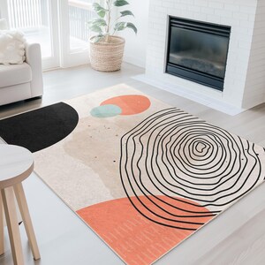 May include: An abstract area rug with a beige background and geometric shapes. The rug features a black semi-circle, a coral-colored rectangle, and a circular design with black lines. Additional shapes include a coral circle and a light blue circle.