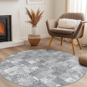 May include: A round, gray area rug with a distressed, geometric pattern. The rug is placed on a hardwood floor, with a chair, side table, and fireplace in the background. The rug's design features a mix of squares and rectangles.