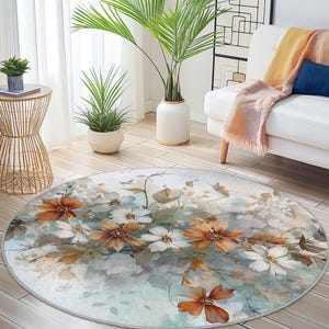May include: A circular rug with a floral design in watercolour style. The rug features orange, white, and blue flowers against a muted background. It has a light grey border and is placed on a light wood floor.