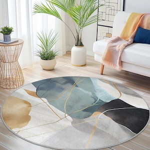 Può includere: Tappeto rotondo con un design astratto nei toni del blu, grigio e oro. Il tappeto presenta forme simili ad acquerelli e linee dorate, creando un aspetto moderno e artistico. Il tappeto è posizionato su un pavimento in legno in un soggiorno.