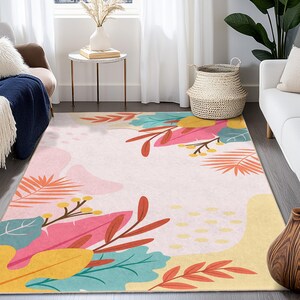 May include: A rectangular area rug with a light pink background and a colorful floral design. The rug features large leaves in shades of pink, yellow, teal, and orange. The rug is placed in a living room setting.