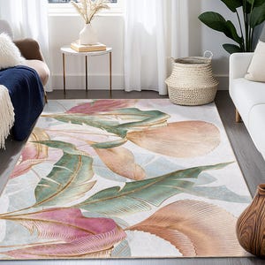 Puede incluir: Una alfombra rectangular con un diseño botánico. La alfombra presenta grandes hojas estilizadas en tonos rosa, verde y melocotón con detalles dorados, sobre un fondo azul claro y blanco. La alfombra está colocada en un salón.