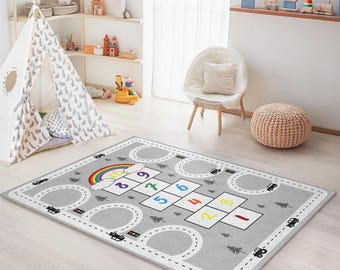 May include: A rectangular grey children's play rug with a hopscotch grid and road design. The rug features a rainbow, clouds, and numbers 1-10 in various colours. Black cars and trees are also depicted around the roads.