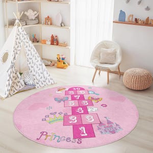 May include: A pink, round hopscotch rug with a princess theme. The rug features a hopscotch grid with numbers, butterflies, a crown, and a castle. The word "Princess" is written in cursive at the bottom.