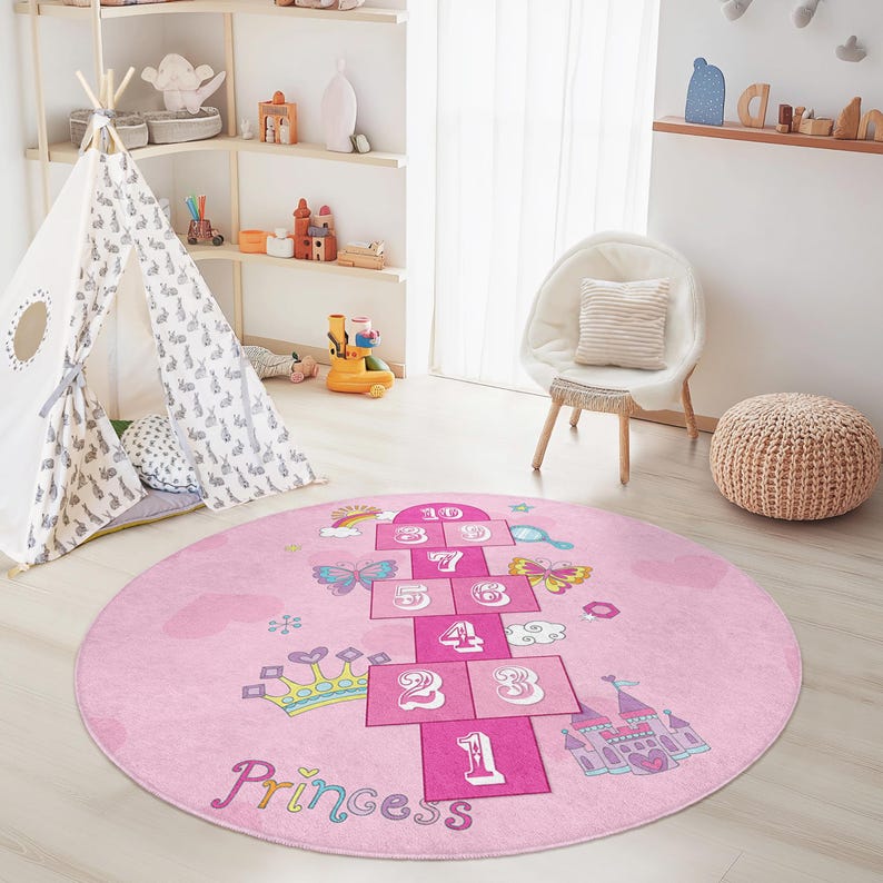 May include: A pink, round hopscotch rug with a princess theme. The rug features a hopscotch grid with numbers, butterflies, a crown, and a castle. The word "Princess" is written in cursive at the bottom.