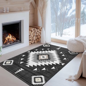 K&ouml;nnte beinhalten: Ein dunkelgrauer Teppich mit einem geometrischen Muster in Wei&szlig;, Grau und Beige. Der Teppich hat ein zentrales Rautenmuster und sich wiederholende geometrische Formen an den R&auml;ndern. Der Teppich liegt auf einem hellen Holzboden vor einem Kamin.