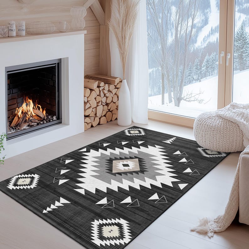 K&ouml;nnte beinhalten: Ein dunkelgrauer Teppich mit einem geometrischen Muster in Wei&szlig;, Grau und Beige. Der Teppich hat ein zentrales Rautenmuster und sich wiederholende geometrische Formen an den R&auml;ndern. Der Teppich liegt auf einem hellen Holzboden vor einem Kamin.