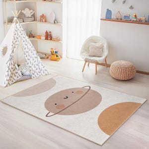 Könnte beinhalten: Rechteckiger Teppich mit Planetenmuster in einem Kinderzimmer. Der Teppich zeigt einen lächelnden Planeten mit einem Ring in Braun- und Beigetönen auf cremefarbenem Hintergrund. Zusätzliche geometrische Formen in ähnlichen Farben ergänzen das Design.