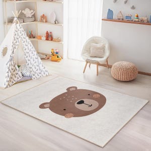 Puede incluir: Alfombra rectangular con una gran cara de oso de dibujos animados en tonos marrón y beige. La alfombra está colocada sobre un suelo de madera clara en una sala de juegos para niños. Una tienda tipi blanca y una silla son visibles en el fondo.