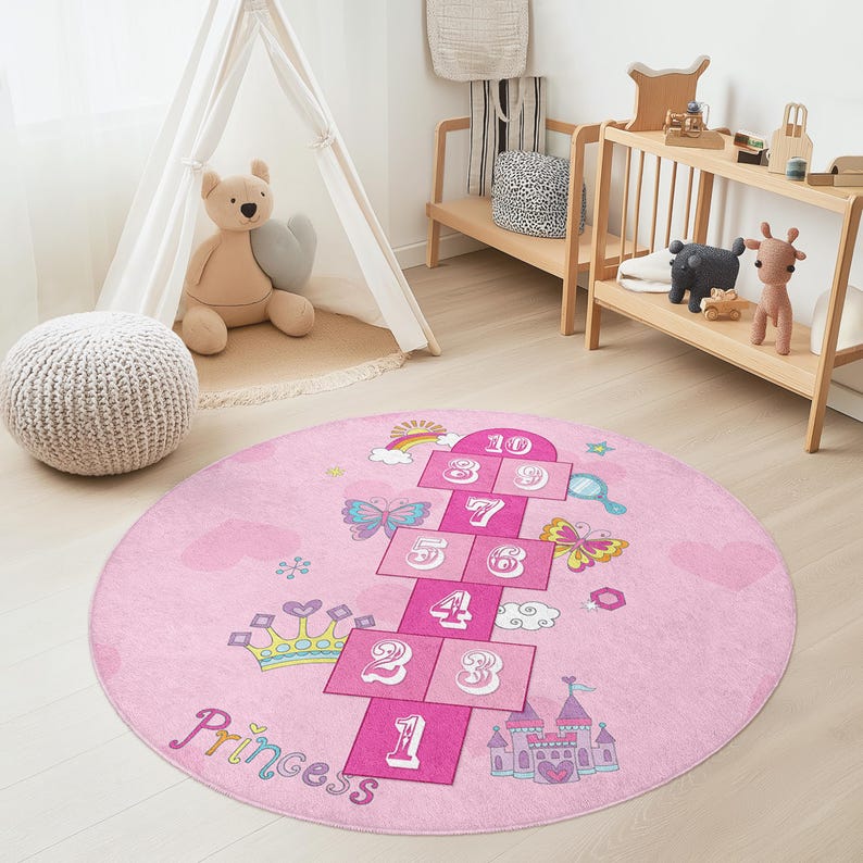 May include: A round pink hopscotch rug with a princess theme. The rug features a hopscotch grid with numbers, butterflies, a rainbow, a crown, and a castle. The word "Princess" is written in cursive.