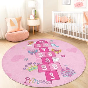 May include: A pink, round hopscotch rug with a princess theme. The rug features a hopscotch grid with numbers, butterflies, a rainbow, a crown, and a castle. The word "Princess" is written at the bottom.