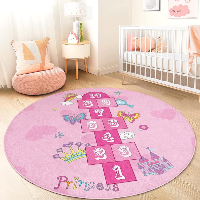 May include: A pink, round hopscotch rug with a princess theme. The rug features a hopscotch grid with numbers, butterflies, a rainbow, a crown, and a castle. The word "Princess" is written at the bottom.