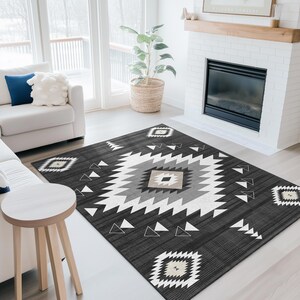 K&ouml;nnte beinhalten: Ein dunkelgrauer Teppich mit einem geometrischen Muster in Wei&szlig;, Hellgrau und Beige. Der Teppich hat ein zentrales Rautenmuster und sich wiederholende dreieckige und rautenf&ouml;rmige Formen. Der Teppich befindet sich in einem Wohnzimmer mit wei&szlig;en M&ouml;beln und einem Kamin.
