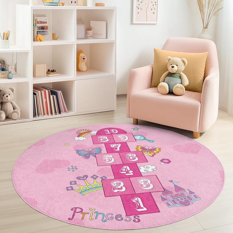 May include: A round pink rug with a hopscotch design. The rug features a hopscotch grid with numbers, butterflies, a crown, and a castle. The word "Princess" is written at the bottom. The rug is in a child's room.