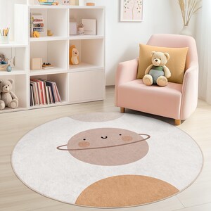Puede incluir: Una alfombra redonda de color blanquecino con un dise&ntilde;o de planeta de dibujos animados en tonos marrones y beige. El planeta tiene una cara sonriente y un anillo. La alfombra est&aacute; en la habitaci&oacute;n de un ni&ntilde;o con un sill&oacute;n rosa y una estanter&iacute;a blanca.