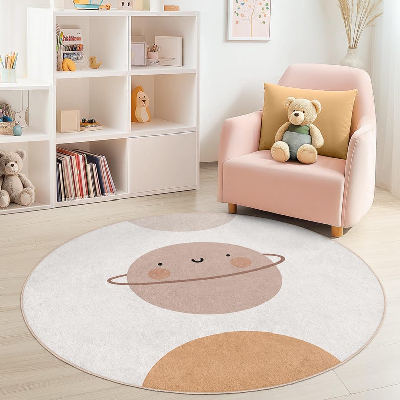 Puede incluir: Una alfombra redonda de color blanquecino con un dise&ntilde;o de planeta de dibujos animados en tonos marrones y beige. El planeta tiene una cara sonriente y un anillo. La alfombra est&aacute; en la habitaci&oacute;n de un ni&ntilde;o con un sill&oacute;n rosa y una estanter&iacute;a blanca.