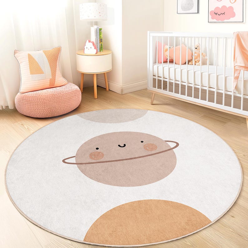 Puede incluir: Una alfombra redonda para ni&ntilde;os, blanca, con un dise&ntilde;o de planeta de dibujos animados. El planeta es de color marr&oacute;n claro con una cara sonriente y un anillo marr&oacute;n. Formas adicionales de color marr&oacute;n claro y gris est&aacute;n en la alfombra.
