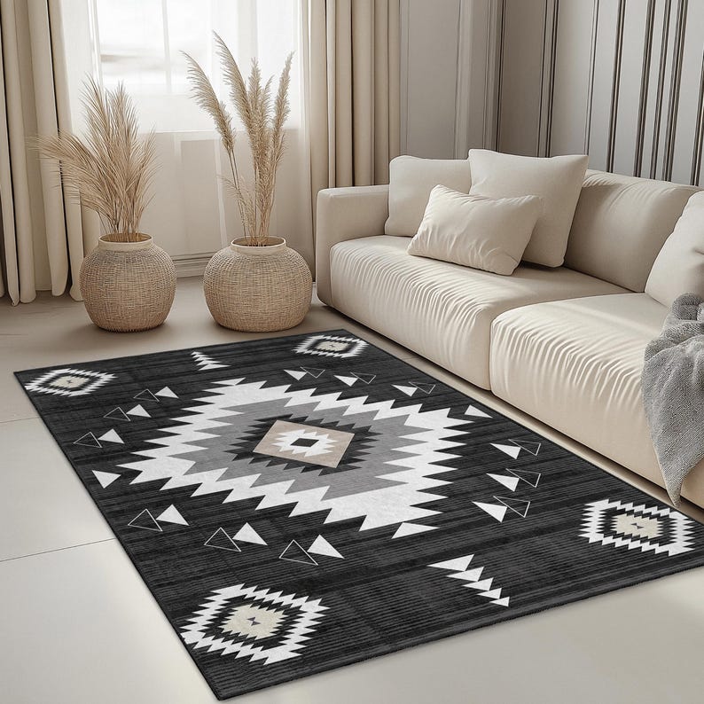 K&ouml;nnte beinhalten: Ein rechteckiger Teppich mit einem geometrischen Muster in Schwarz, Wei&szlig;, Grau und Beige. Der Teppich hat ein zentrales Rautenmuster, das von Dreiecken und Rautenformen umgeben ist. Der Teppich liegt auf einem hellen Boden.