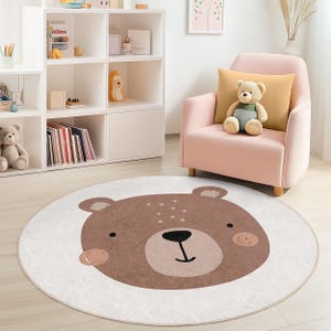 Puede incluir: Una alfombra redonda y blanca con un diseño de cara de oso marrón grande. El oso tiene un hocico marrón claro, ojos oscuros y una boca negra. La alfombra está en la habitación de un niño con un sillón rosa y un oso de peluche.