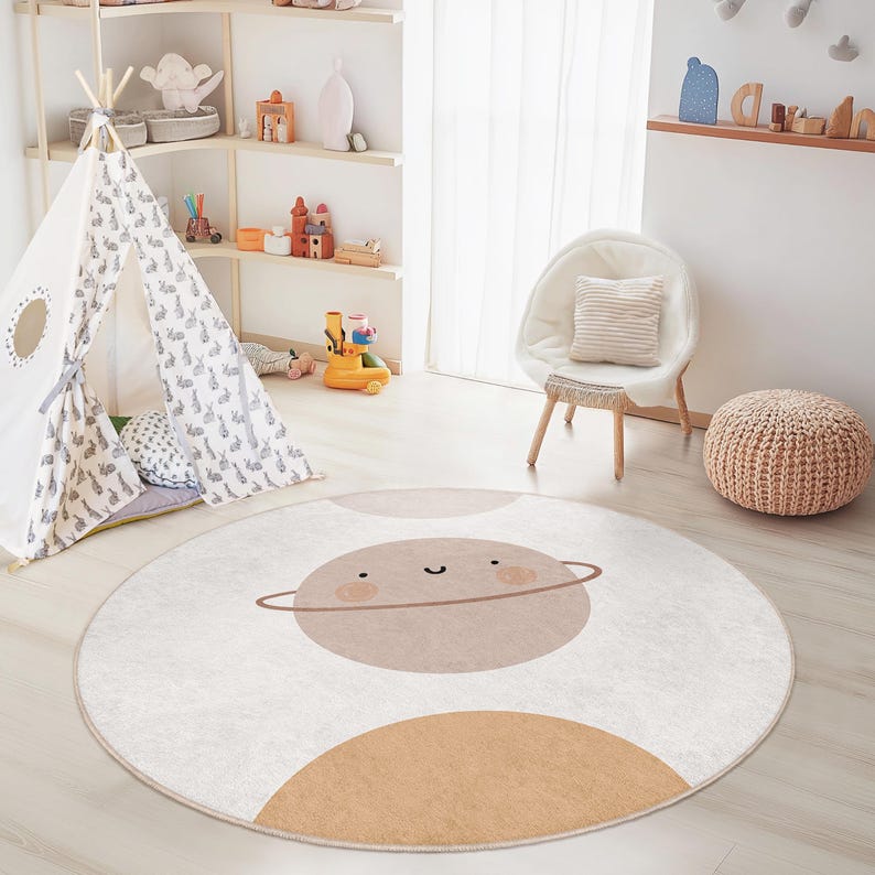 Puede incluir: Alfombra redonda infantil, de color blanco roto, con dise&ntilde;o de planeta. El planeta es marr&oacute;n claro con una cara sonriente y un anillo marr&oacute;n. Una gran semicircunferencia marr&oacute;n claro est&aacute; en la parte inferior. La alfombra est&aacute; en un cuarto infantil.